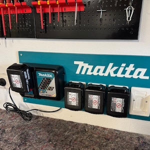 Könnte beinhalten: Eine türkisfarbene Makita-Ladestation mit weißem Makita-Logo. Die Station enthält mehrere schwarze Akkus und ein weißes Zubehörteil. Ein schwarzes Netzkabel ist an eine Steckdose angeschlossen. Rote und gelbe Werkzeuge hängen darüber.