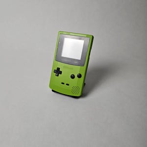 Könnte beinhalten: Eine limettengrüne Game Boy Color Handheld-Spielkonsole mit einem schwarzen Steuerkreuz, zwei schwarzen Aktionstasten, einer Start-Taste, einer Select-Taste, einem Lautsprechergitter und einem Bildschirm mit dem Nintendo Game Boy Color Logo.