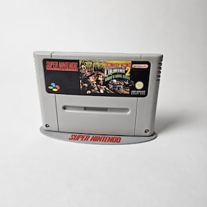 Könnte beinhalten: Ein graues Super Nintendo Videospielmodul für Donkey Kong Country 2 Diddy's Kong Quest. Das Etikett zeigt den Spieletitel, das Nintendo-Logo und Charaktergrafiken. Das Modul steht auf einem grauen, ovalen Ständer mit dem Super Nintendo-Logo.