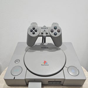 Könnte beinhalten: Eine graue Sony PlayStation-Konsole mit passendem Controller. Die Konsole verfügt über ein Laufwerk, Ein- und Aus-Tasten sowie das PlayStation-Logo. Der Controller hat Richtungstasten, Aktionstasten und eine Starttaste.