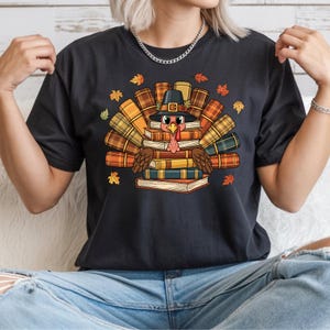 May include: Black t-shirt featuring a cartoon turkey wearing a pilgrim hat and surrounded by books. The turkey is in the center of the shirt, with books forming a fan shape around it. Autumn leaves are scattered around the turkey.
