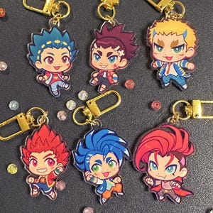 May include: A collection of colorful anime-style character keychains. Each keychain features a unique character design with vibrant hair and clothing, attached to a gold-colored clasp. The keychains are arranged on a dark surface with scattered decorative gems.