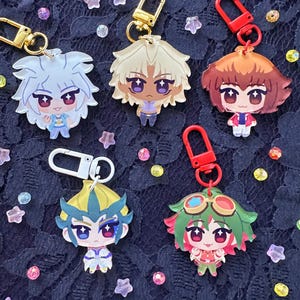 May include: Five anime-style character keyrings with gold and red clasps. The keyrings feature colourful, detailed illustrations of characters with various hair colours and outfits. The background is a dark, textured fabric with scattered decorative beads.