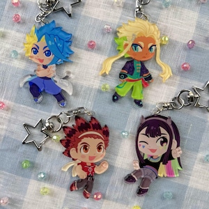 Può includere: Quattro portachiavi con personaggi in stile anime. Ogni portachiavi presenta un personaggio diverso con colori unici di capelli e abbigliamento. I portachiavi sono attaccati a ciondoli a forma di stella e portachiavi color argento. Lo sfondo è un motivo a scacchi blu chiaro e bianco.