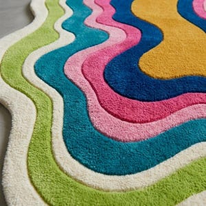 Colorful wavy abstract area rug featuring layered organic curves in green, turquoise, pink, navy blue, mustard yellow, and ivory. Plush raised tufted texture and sculptural irregular shape create a bold modern statement. Shown in a luxury hallway and on a neutral floor, this colorful area rug is ideal for living rooms, bedrooms, hallways, studios, offices, and contemporary interiors.