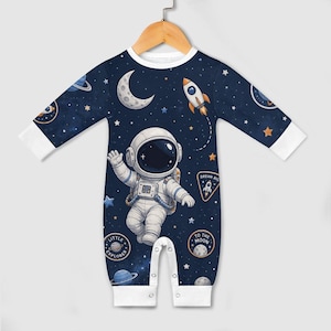 Op de afbeelding: Marineblau baby-onesie met een astronaut, sterren, planeten en een maan. De onesie met lange mouwen heeft witte manchetten en een witte halslijn. Patches met de tekst "Little Explorer", "To the Moon" en "Dream Big".