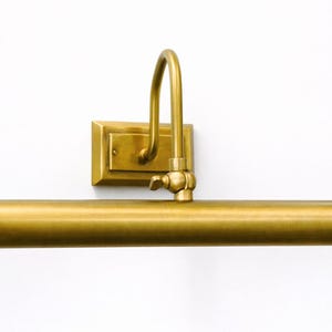 May include: A wall-mounted picture light with a brushed brass finish. The fixture features a rectangular backplate, a curved arm, and a horizontal cylindrical light bar. A small adjustable knob is located at the joint connecting the arm to the bar.