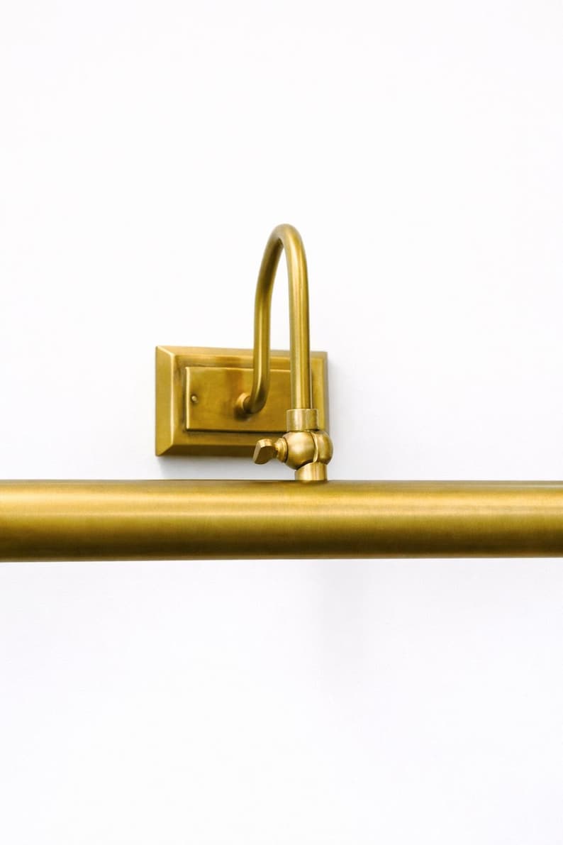 May include: A wall-mounted picture light with a brushed brass finish. The fixture features a rectangular backplate, a curved arm, and a horizontal cylindrical light bar. A small adjustable knob is located at the joint connecting the arm to the bar.