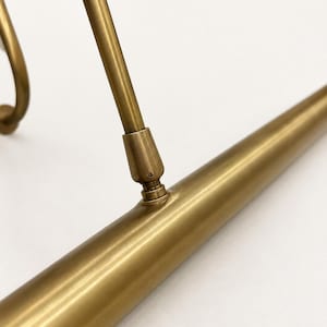 May include: A brass picture light fixture featuring a long, cylindrical horizontal tube connected to a slender, angled support arm. The metal has a smooth, matte finish with visible joints where the arm attaches to the main light housing.