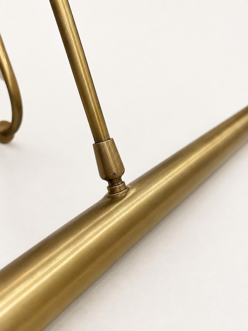 May include: A brass picture light fixture featuring a long, cylindrical horizontal tube connected to a slender, angled support arm. The metal has a smooth, matte finish with visible joints where the arm attaches to the main light housing.