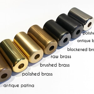 May include: A set of seven cylindrical metal hardware components arranged in a diagonal row. The finishes include antique patina, polished brass, brushed brass, raw brass, blackened brass, antique black, and polished chrome, each with a center hole.