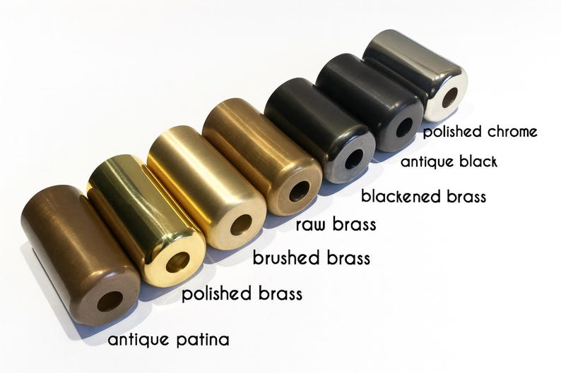 May include: A set of seven cylindrical metal hardware components arranged in a diagonal row. The finishes include antique patina, polished brass, brushed brass, raw brass, blackened brass, antique black, and polished chrome, each with a center hole.