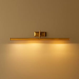 May include: A horizontal wall-mounted picture light fixture with a metallic brass finish. The design features a long, slender cylindrical tube attached to a rectangular base plate by a central vertical arm, emitting a warm, downward glow.