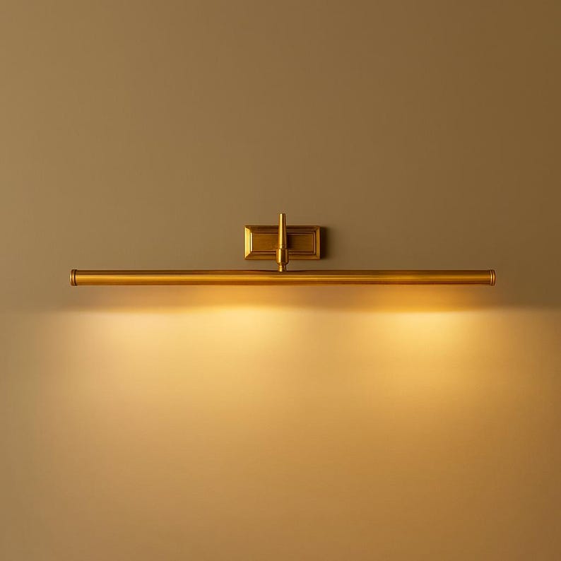 May include: A horizontal wall-mounted picture light fixture with a metallic brass finish. The design features a long, slender cylindrical tube attached to a rectangular base plate by a central vertical arm, emitting a warm, downward glow.