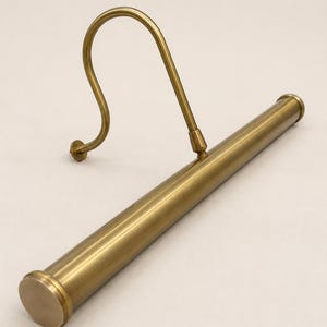 May include: A brass-toned metal picture light featuring a long, cylindrical horizontal tube attached to a curved, S-shaped mounting arm. The fixture has a smooth, metallic finish with circular end caps on the tube and a small circular wall plate.