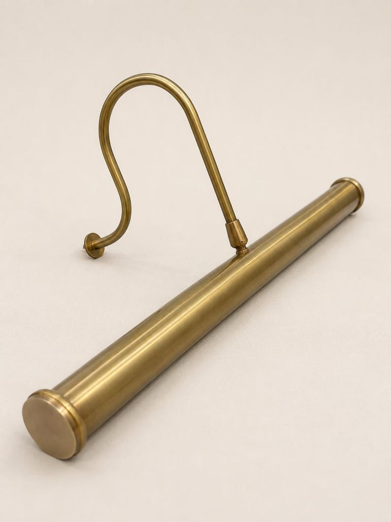 May include: A brass-toned metal picture light featuring a long, cylindrical horizontal tube attached to a curved, S-shaped mounting arm. The fixture has a smooth, metallic finish with circular end caps on the tube and a small circular wall plate.