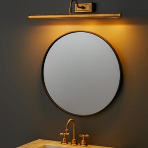 May include: A round mirror with a thin dark frame mounted above a white marble vanity top with a brass faucet. A horizontal brass picture light is installed on the wall directly above the mirror, casting a warm glow downward onto the glass surface.