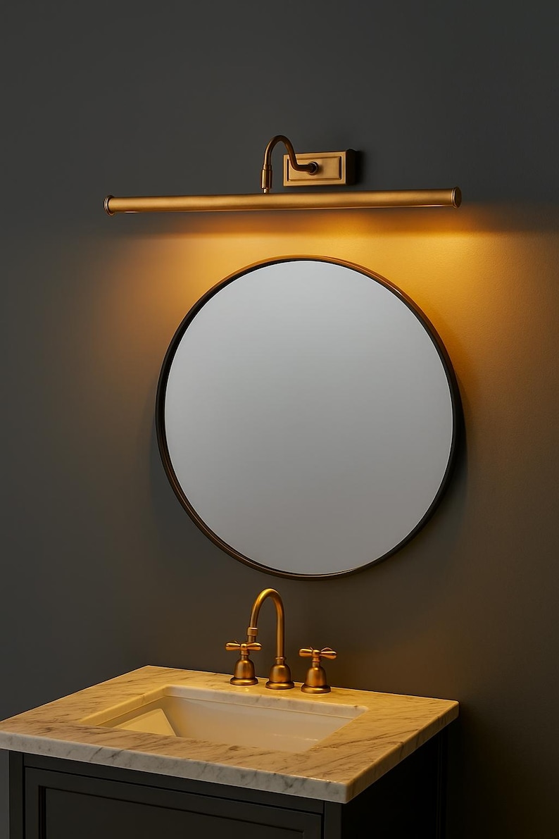 May include: A round mirror with a thin dark frame mounted above a white marble vanity top with a brass faucet. A horizontal brass picture light is installed on the wall directly above the mirror, casting a warm glow downward onto the glass surface.