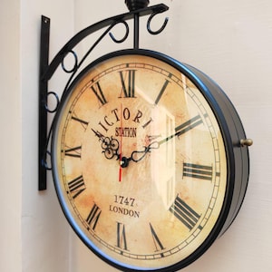 Puede incluir: Un reloj de pared de metal negro de doble cara con un diseño vintage. La esfera del reloj es de color crema con números romanos negros y las palabras "VICTORIA STATION 1747 LONDON". El reloj tiene un segundero rojo.