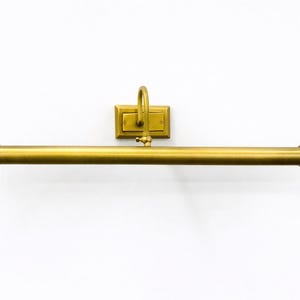 May include: A wall-mounted picture light with a long, cylindrical horizontal bar and a rectangular backplate, finished in a brushed brass tone. The fixture features a curved arm connecting the central bar to the mounting plate.