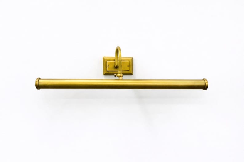 May include: A wall-mounted picture light with a long, cylindrical horizontal bar and a rectangular backplate, finished in a brushed brass tone. The fixture features a curved arm connecting the central bar to the mounting plate.