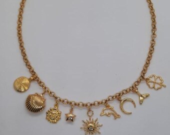 May include: Gold-toned charm necklace featuring various pendants, including a seashell, sun, star, dolphin, and clover. The necklace has a delicate chain and is displayed against a white background. The charms are detailed and add a touch of whimsy.