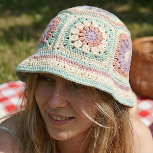 May include: A crocheted bucket hat in pastel colors, featuring a floral pattern. The hat's design incorporates squares of light blue, cream, pink, and purple. The hat is worn outdoors, with a picnic basket and bowl of berries in the background.