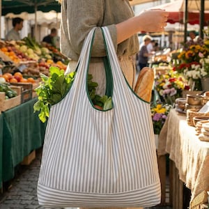 Puede incluir: Un bolso tote a rayas con ribete verde, lleno de productos frescos y una baguette, se lleva en un mercado al aire libre. La bolsa presenta rayas verticales grises y blancas y la lleva una persona. El mercado tiene varias frutas, verduras y flores.