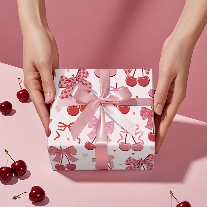 May include: A wrapped gift with a white background featuring a pattern of red cherries and pink bows. The gift is tied with a pink satin ribbon and bow. Fresh cherries are scattered around the gift on a pink surface.