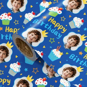 May include: Blue birthday wrapping paper with a pattern of cupcakes, stars, crowns, and a smiling child's face. The words "Happy Birthday" are written in yellow. A roll of the wrapping paper is visible.