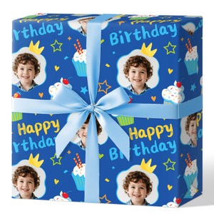 May include: A wrapped gift box with a blue ribbon. The wrapping paper is blue and features a repeating pattern of a child's face, cupcakes, crowns, and the words "Happy Birthday".