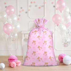 Pode incluir: Uma grande sacola de presente rosa com um padrão repetido de ursinhos de pelúcia marrons e laços rosa. A sacola é amarrada no topo com uma fita rosa. Balões rosa e brancos e decorações de papel no fundo.