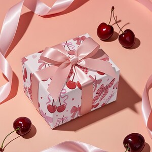 May include: A wrapped gift box with a pink satin bow and ribbon. The box is white with a pattern of red cherries and pink bows. Fresh cherries are scattered around the box on a pink background.