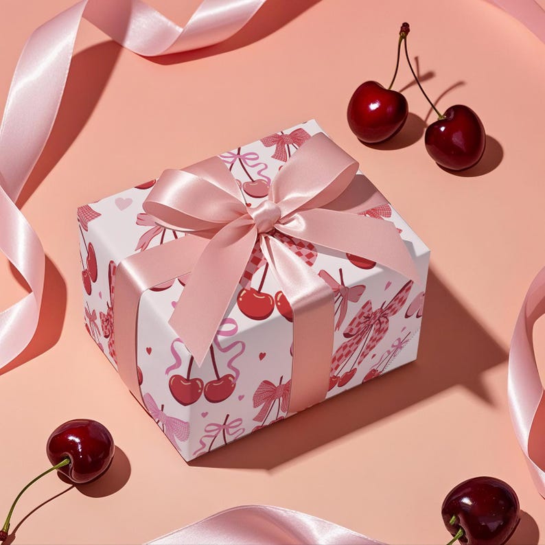 May include: A wrapped gift box with a pink satin bow and ribbon. The box is white with a pattern of red cherries and pink bows. Fresh cherries are scattered around the box on a pink background.