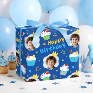 May include: A wrapped gift box with a blue ribbon, featuring a pattern of cupcakes, stars, crowns, and a child's photo on a dark blue background. The words "Happy Birthday" are also visible. Balloons and cupcakes are in the background.