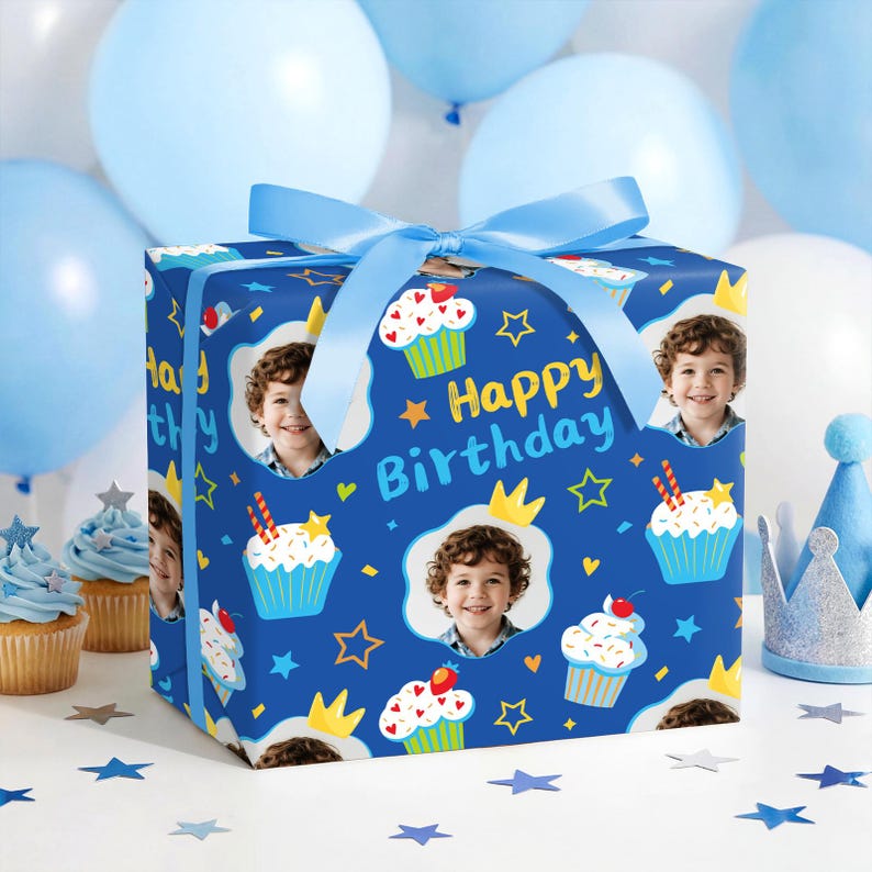 May include: A wrapped gift box with a blue ribbon, featuring a pattern of cupcakes, stars, crowns, and a child's photo on a dark blue background. The words "Happy Birthday" are also visible. Balloons and cupcakes are in the background.