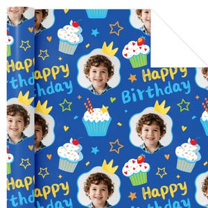 May include: Blue wrapping paper with a repeating pattern of cupcakes, stars, crowns, and the text "Happy Birthday." The design includes a photo of a smiling child. The cupcakes have white frosting and red cherries.