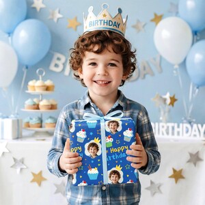 May include: A young child wearing a crown holds a wrapped gift. The gift wrap is blue with cupcakes, stars, and the child's photo. The background includes balloons, cupcakes, and a banner that says "Birthday".