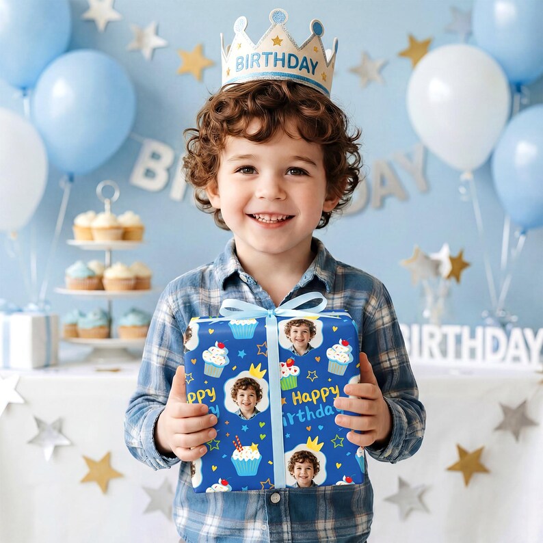 May include: A young child wearing a crown holds a wrapped gift. The gift wrap is blue with cupcakes, stars, and the child's photo. The background includes balloons, cupcakes, and a banner that says "Birthday".