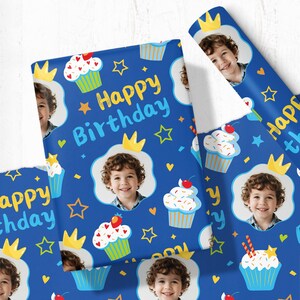 May include: Blue birthday wrapping paper with a pattern of cupcakes, crowns, stars, and a child's photo. The words "Happy Birthday" are written in yellow and blue. The paper is rolled and flat.