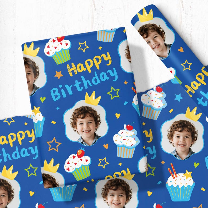 May include: Blue birthday wrapping paper with a pattern of cupcakes, crowns, stars, and a child's photo. The words "Happy Birthday" are written in yellow and blue. The paper is rolled and flat.