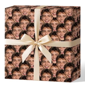 Può includere: Scatola regalo quadrata avvolta in carta con un motivo ripetuto di volti sorridenti. Un nastro e un fiocco color crema adornano il pacco, creando una presentazione decorativa e originale per un regalo.