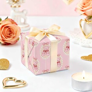 Pode incluir: Uma caixa de presente quadrada embrulhada em papel rosa com um padr&atilde;o repetido de ursinhos em molduras ovais. &Eacute; amarrada com uma fita e la&ccedil;o creme. O fundo inclui rosas, uma vela e decora&ccedil;&otilde;es douradas em forma de cora&ccedil;&atilde;o.