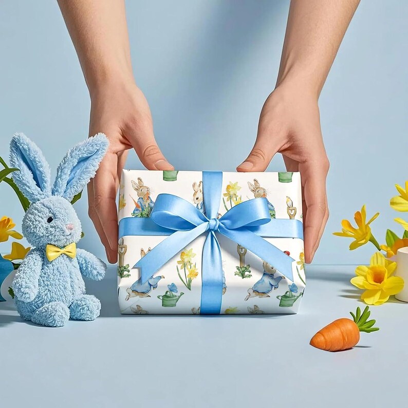 May include: A wrapped gift with a blue ribbon, featuring a pattern of bunnies and daffodils. A plush blue bunny with a yellow bow tie sits beside the gift. A small orange carrot and yellow flowers complete the Easter-themed scene.