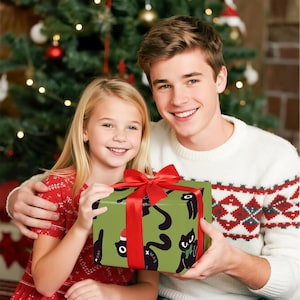 May include: A wrapped gift with a red bow is held by two people. The gift is wrapped in green paper with a black cat pattern. The people are in front of a Christmas tree. One person is wearing a white sweater with a red and gray pattern.