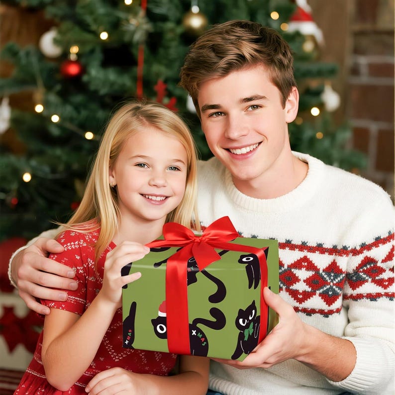 May include: A wrapped gift with a red bow is held by two people. The gift is wrapped in green paper with a black cat pattern. The people are in front of a Christmas tree. One person is wearing a white sweater with a red and gray pattern.