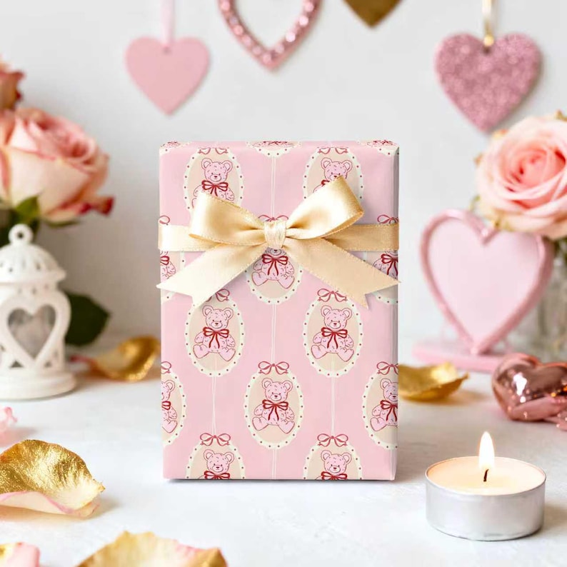 Pode incluir: Uma caixa de presente embrulhada em papel rosa com um padr&atilde;o repetido de ursinhos de pel&uacute;cia em molduras ovais. Uma fita dourada est&aacute; amarrada ao redor da caixa. O fundo inclui cora&ccedil;&otilde;es rosa, rosas e uma vela acesa.