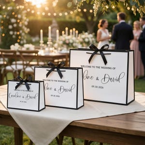 May include: Three white gift bags with black trim and black ribbon handles are displayed on a table. The bags have the text "WELCOME TO THE WEDDING OF" and the names "Caroline & David" and "Line & David" with the date "09.30.2026" or "09.30.2026".