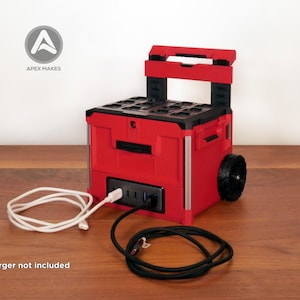 Puede incluir: Una estación de energía portátil roja y negra con un asa superior y ruedas. La unidad cuenta con un panel frontal con múltiples tomas de corriente y puertos USB. Dos cables de carga están conectados. El texto APEX MAKES es visible.