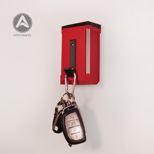 Puede incluir: Un soporte de pared para llaves rojo y negro con cuerpo rectangular y gancho negro. Un mosquetón plateado con un juego de llaves y un mando de coche cuelga del gancho. El logotipo de APEX MAKES es visible en la esquina superior izquierda.